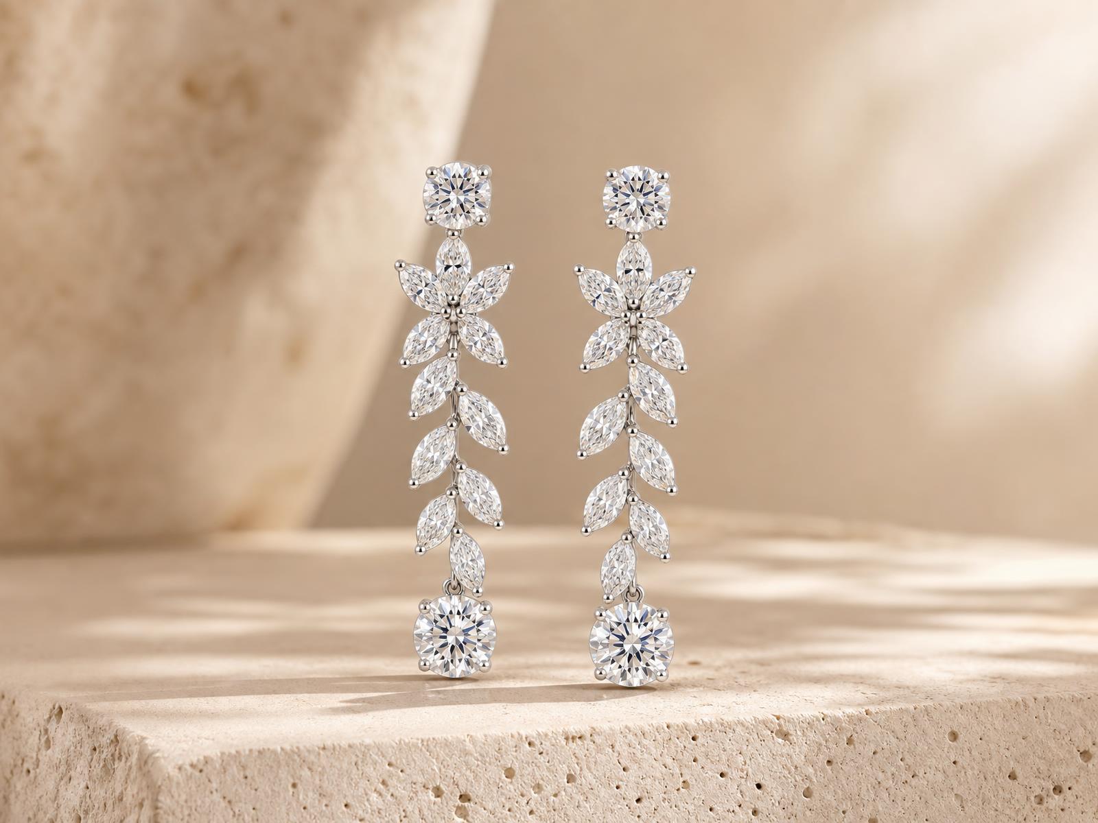 Pair of RW floral vine diamond drop earrings in 14k white gold, round and marquise diamonds, on a warm cream textured concrete ledge