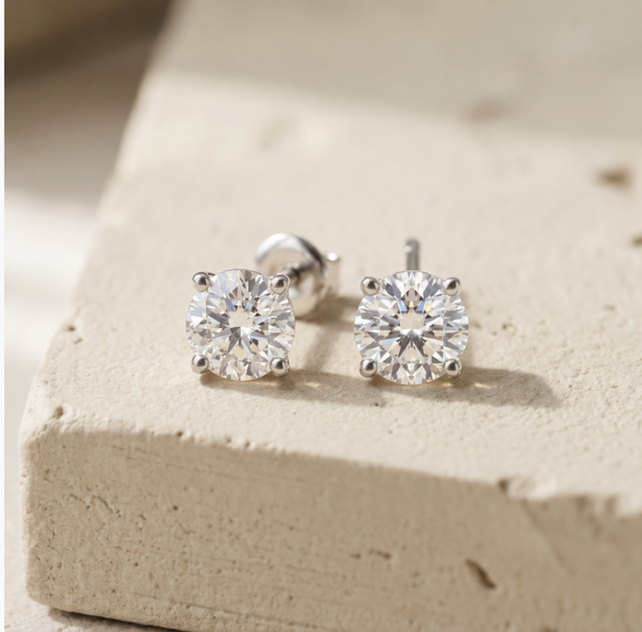 Pair of RW round brilliant diamond studs in a low-profile four-claw flower-style setting on warm cream concrete